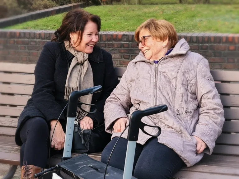 Seniorin und Betreuerin sitzen lachend auf einer Parkbank – herzliche Begleitung durch Mia Oppermann.