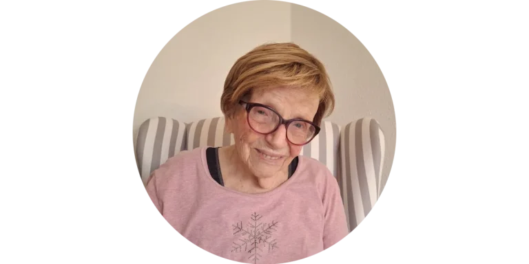 Lächelnde ältere Dame mit Brille und oranger Strickjacke sitzt entspannt auf dem Sofa – Kundin von Mia Oppermann.