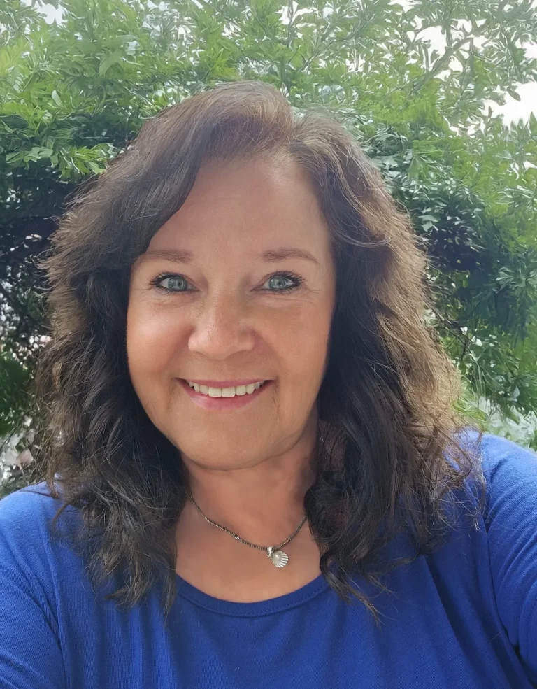 Frau mit lockigem, braunem Haar trägt ein blaues Shirt und lächelt vor grüner Natur.