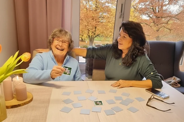 Seniorin und Betreuerin lachen beim Memoryspiel am Tisch – herzliche Begleitung durch Mia Oppermann.